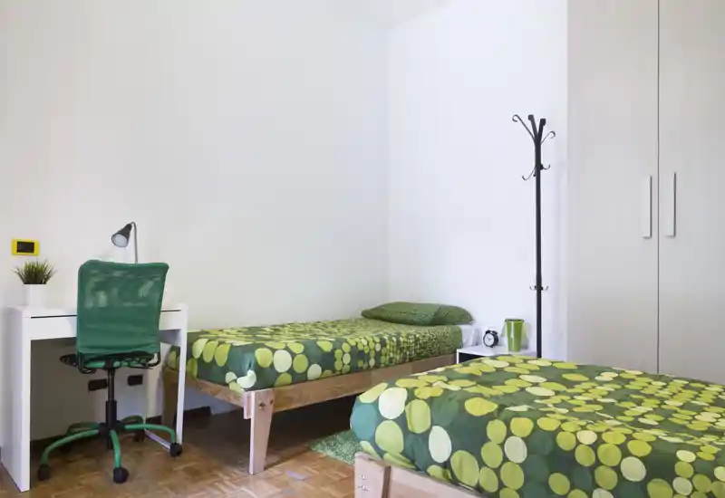 Chambre twin lumineuse avec deux lits simples habillés d'une literie verte coordonnée, petit bureau blanc et chaise, armoire et décoration épurée — propre et prête à accueillir.