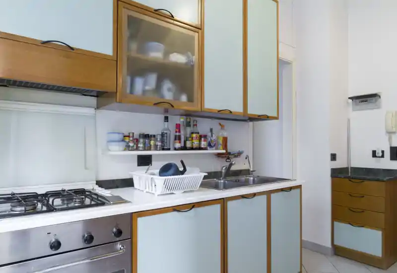 Compact fitted kitchen with gas hob, stainless oven, double sink and upper cabinets; spices and a dish rack indicate active use and practical storage.