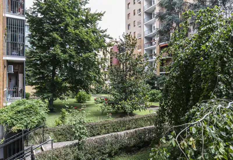 Communal garden courtyard framed by mature trees and hedges, with lawn beds and surrounding apartment balconies — a pleasant green space for residents.