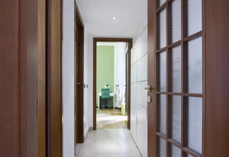 Bright, clean hallway with wooden doors and built-in storage leading to a small study with a green accent wall; well-lit corridor ideal for apartment listings.