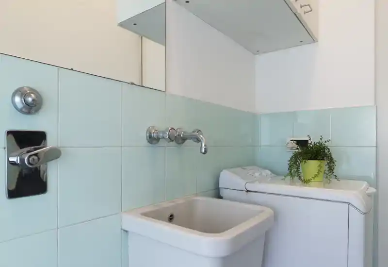 Compact laundry/utility corner with a deep sink, wall-mounted faucet and top-loading washing machine; pale aqua tiles and a small potted plant create a fresh, clean look.