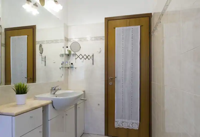 Clean, well-lit bathroom with a vanity sink, mirror, and wooden door — bright tiles and organized countertop suitable for rental listing.