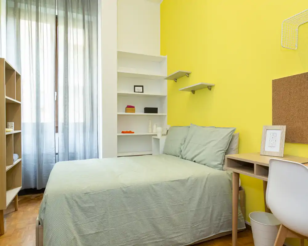 Camera da letto luminosa e ordinata con letto matrimoniale contro una parete gialla vivace, scaffalatura integrata, scrivania e luce naturale proveniente da una finestra con tende leggere.