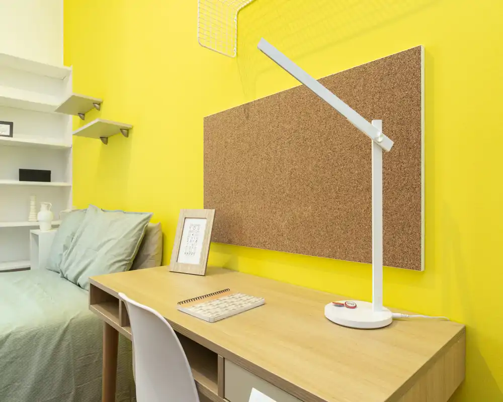 Angolo camera da letto con scrivania in legno, pannello in sughero e letto singolo con biancheria verde. Parete gialla brillante e lampada da tavolo moderna creano un ambiente funzionale e vivace.