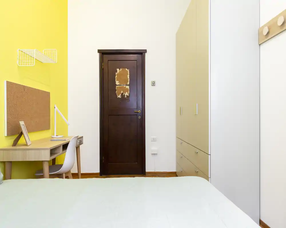 Camera da letto luminosa e ordinata con il letto in primo piano, una porta in legno al centro della parete posteriore, una scrivania con lampada contro una parete gialla e un armadio a destra.