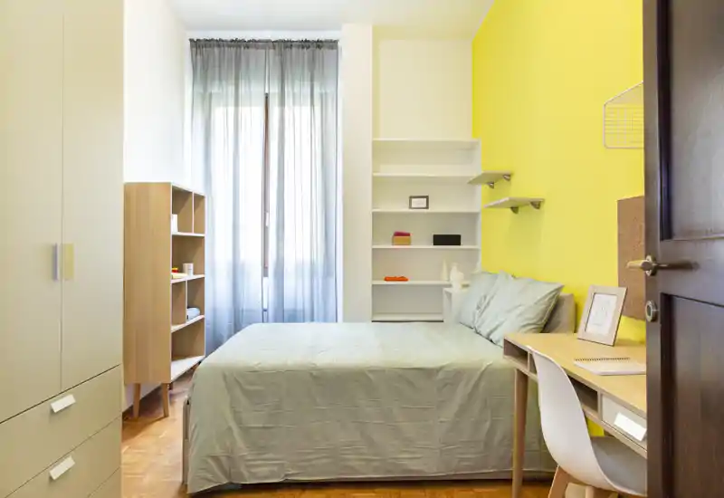 Camera da letto luminosa e accogliente con letto matrimoniale ben rifatto, scrivania in legno, scaffalatura e una grande finestra che illumina la stanza.