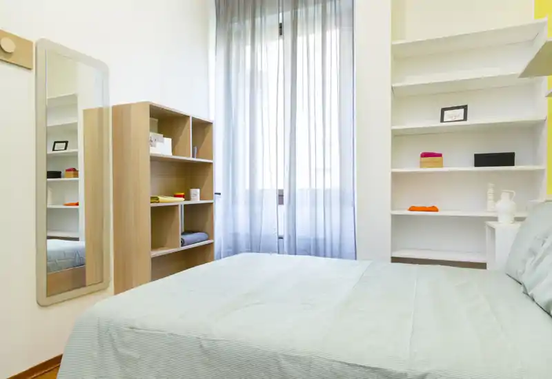 Camera da letto moderna e luminosa con letto ben rifatto, scaffalature aperte e uno specchio alto vicino alla finestra che diffonde luce naturale.