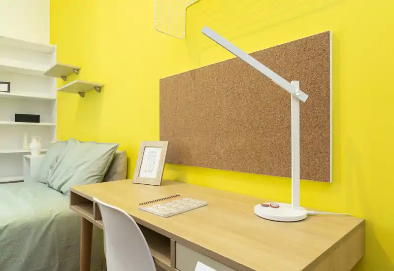 Angolo camera da letto con scrivania in legno, pannello in sughero e letto singolo con biancheria verde. Parete gialla brillante e lampada da tavolo moderna creano un ambiente funzionale e vivace.