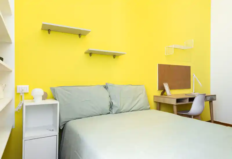 Camera da letto moderna e luminosa con letto matrimoniale ben rifatto davanti a una parete gialla, comodino e una scrivania con sedia — pulita e ben illuminata.