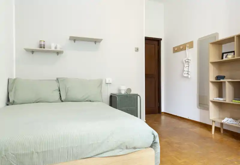 Camera da letto luminosa e minimale con un letto matrimoniale ben rifatto con biancheria verde chiaro, mensole semplici sopra la testata, un comodino e una libreria aperta a lato. La stanza è ariosa e ben illuminata, perfetta per un annuncio in affitto che valorizzi comfort e stile pulito.