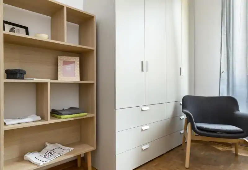 Angolo camera ben illuminato con armadio bianco, scaffalatura in legno a vista e una comoda poltrona imbottita, ideale per riporre oggetti e creare un angolo lettura.
