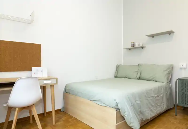 Camera da letto luminosa e minimal con letto matrimoniale con lenzuola verde salvia, piccole mensole e una scrivania compatta — ideale per uno studente o una persona sola.
