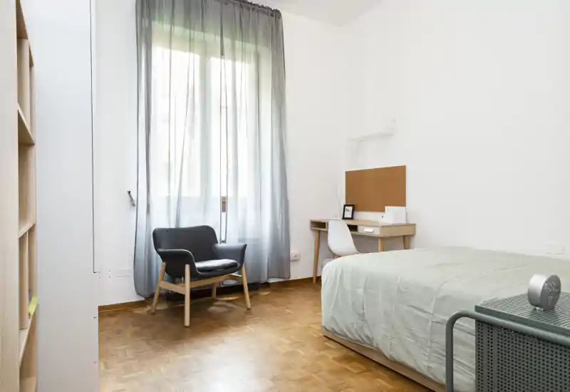 Camera da letto luminosa e minimal con letto singolo, scrivania e una poltrona confortevole accanto a una grande finestra che lascia entrare luce naturale.