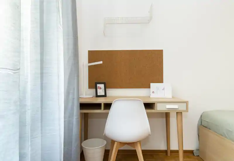 Angolo camera compatto con scrivania in legno ordinata, sedia e letto parzialmente visibile a destra. Stile pulito e minimal con luce naturale soft, adatto a un ambiente studio-notte.