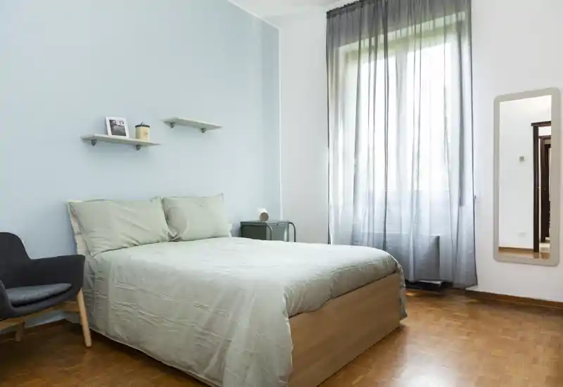 Chambre calme et lumineuse avec lit double, linge de lit vert pâle et décoration scandinave minimaliste ; grande fenêtre avec voilage laissant entrer une belle lumière naturelle.