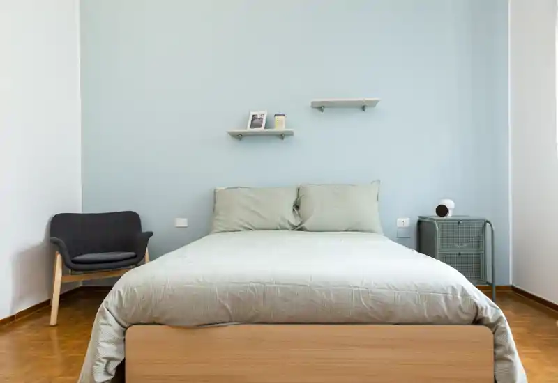 Chambre lumineuse et minimaliste avec un lit double centré, linge de lit pastel et un fauteuil confortable. Des lignes épurées et un mur bleu apaisant créent un espace propice au repos.