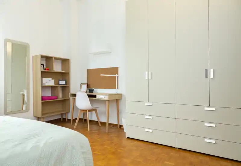 Camera da letto luminosa e minimal con angolo del letto visibile, ampia armadiatura, pavimento in legno e una scrivania compatta — ordinata e ben illuminata per annunci.
