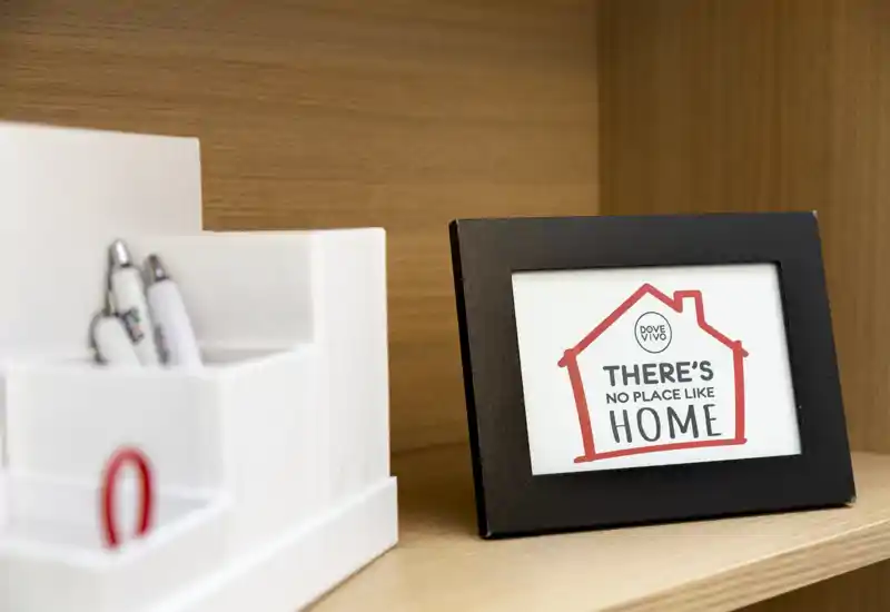 Primo piano di una mensola in legno con una cornice che riporta "There's no place like home" e un organizer bianco con penne; immagine dettagliata di arredo, non una panoramica di una stanza.