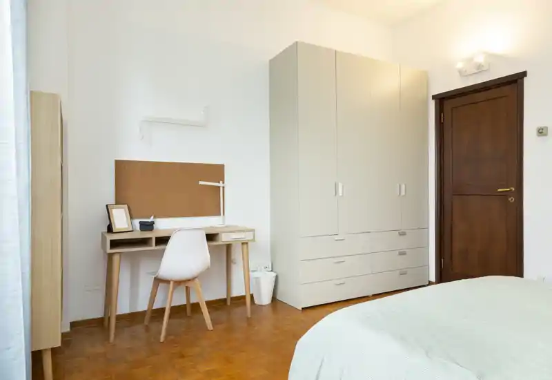 Camera da letto luminosa e minimal con letto visibile in primo piano, armadio alto e una semplice scrivania in legno con sedia — ideale per un annuncio di affitto curato.