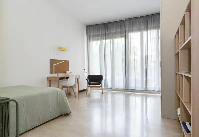 Camera da letto minimalista e luminosa con un letto singolo, una piccola scrivania e una poltrona davanti a grandi finestre con tende leggere che filtrano la luce naturale.