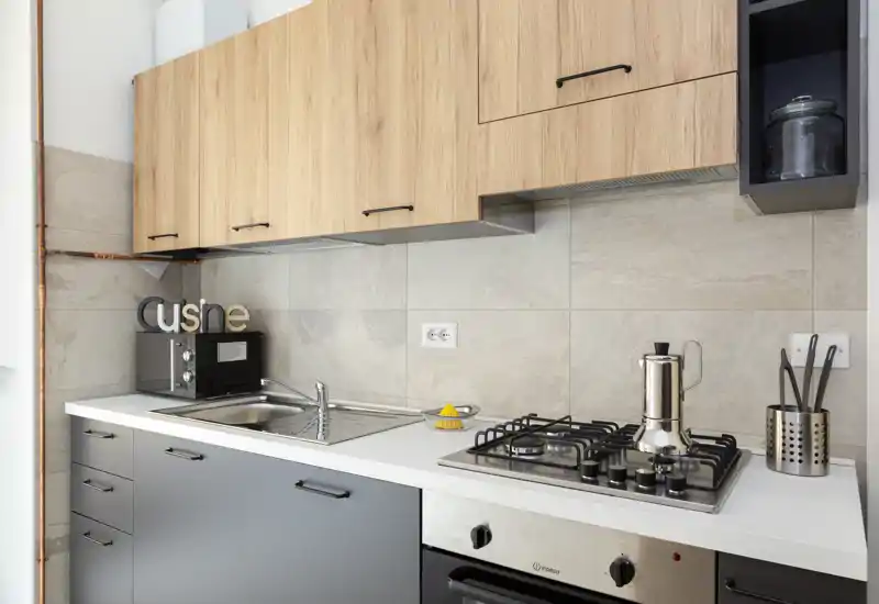 Cocina moderna y compacta con fregadero de acero inoxidable, cocina de gas y armarios superiores de madera — espacio luminoso y bien organizado, ideal para apartamentos urbanos.