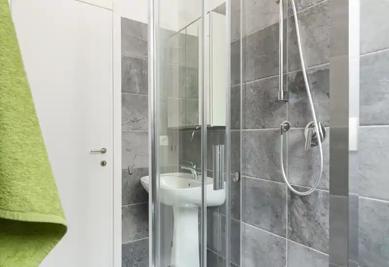 Bagno compatto e moderno con pareti in piastrelle grigie, box doccia angolare in vetro e lavabo a colonna; ambiente luminoso, pulito e funzionale ideale per annunci.