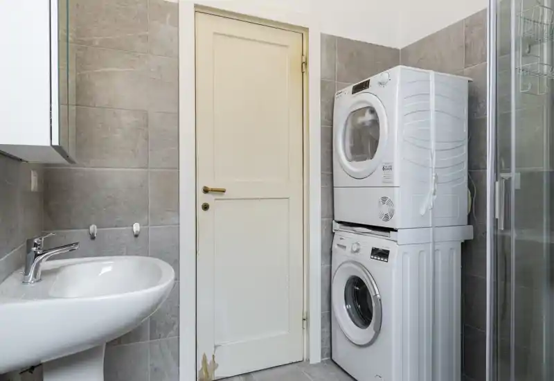 Bagno compatto con lavabo a colonna, lavatrice e asciugatrice impilate e box doccia in vetro; piastrelle grigie neutre e illuminazione pulita creano un ambiente moderno e funzionale.