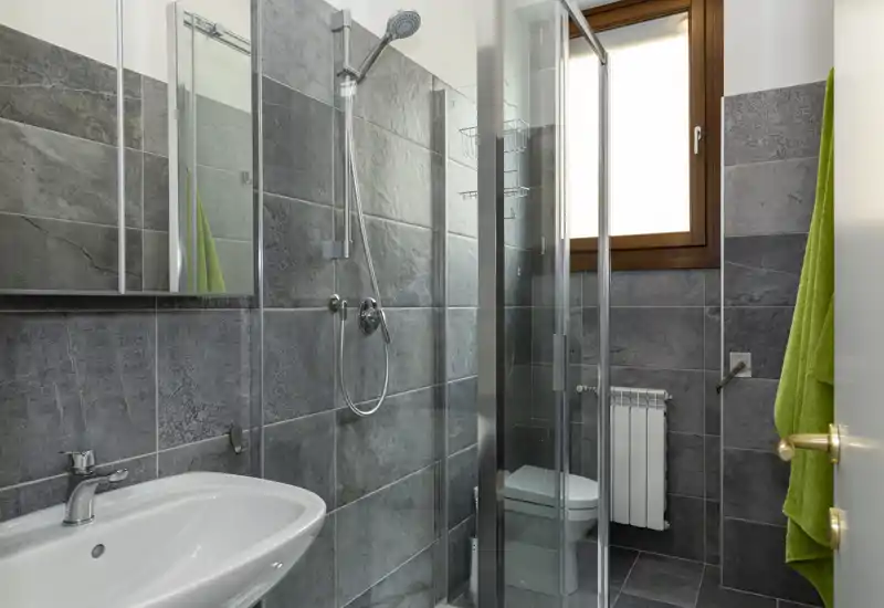 Bagno moderno e compatto con piastrelle grigie, doccia con box in vetro, lavabo a muro e piccolo radiatore sotto la finestra. Layout pulito e funzionale, ideale per affitti o come bagno per gli ospiti.