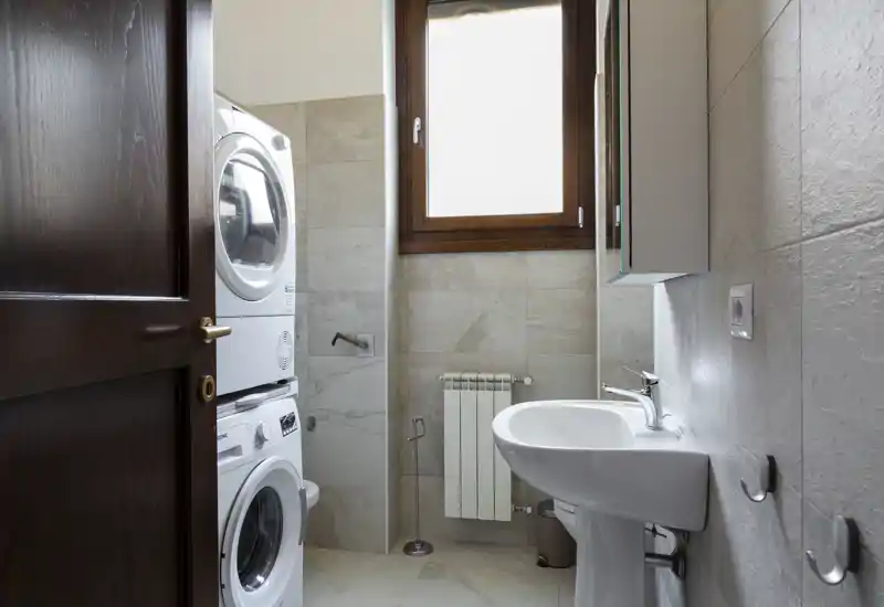 Bagno compatto con lavabo a colonna, pensile specchiato, termosifone e lavatrice asciugatrice impilate accanto alla porta; piastrelle in tonalità neutre e finestra alta con cornice in legno.