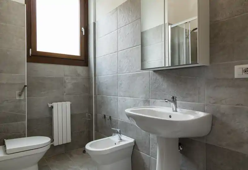 Bagno moderno e ben illuminato con lavabo a colonna, bidet e WC contro grandi piastrelle grigie e un armadietto specchiato sopra il lavabo.