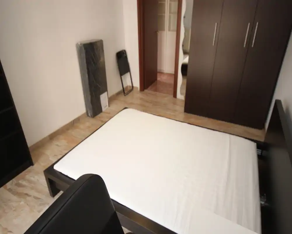 Dormitorio limpio y sencillo con un colchón grande sobre una estructura de cama de madera oscura, armario y suelo de baldosas; tonos neutros y distribución ordenada para un espacio tranquilo.