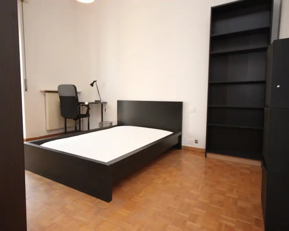 Camera da letto moderna e minimale con struttura letto bassa nera, materasso chiaro e una postazione di lavoro nell'angolo. Pavimento in parquet pulito e scaffalatura alta per riporre oggetti; arredamento essenziale e funzionale.