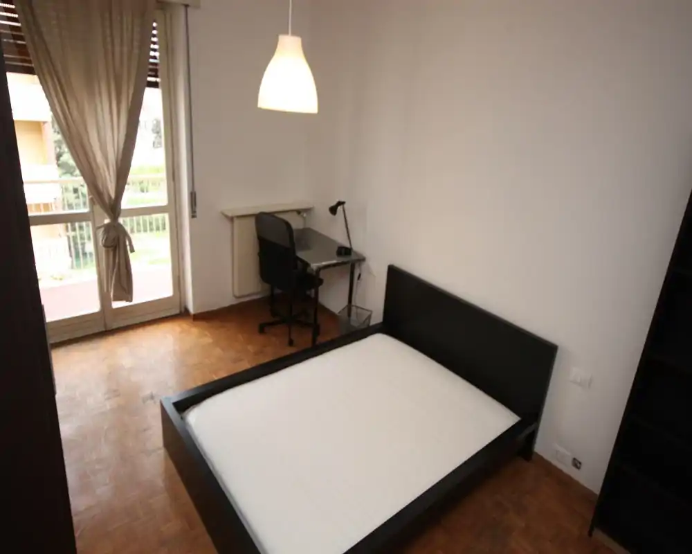 Camera da letto luminosa e minimal con letto matrimoniale e una piccola scrivania vicino al balcone. Pavimento in parquet, lampada a sospensione e porta finestra assicurano luce naturale e un’atmosfera tranquilla.