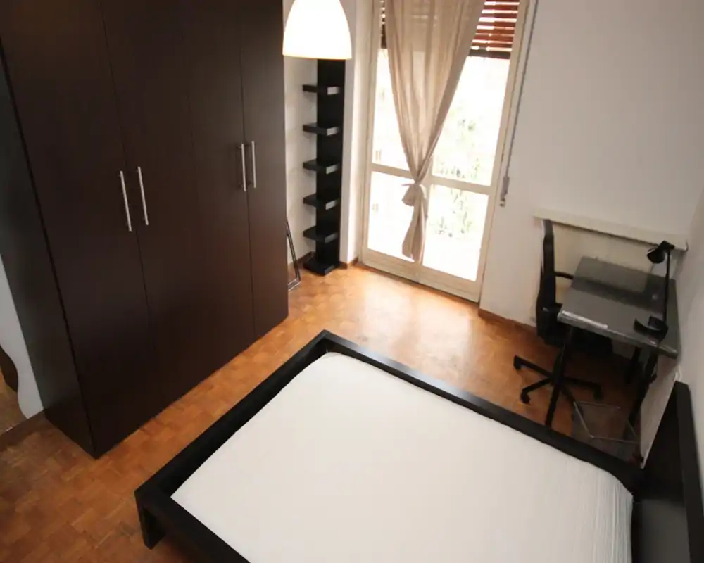 Camera da letto luminosa e moderna con letto matrimoniale su struttura bassa nera, ampio armadio, scrivania vicino alla finestra e pavimento in parquet — ideale per singolo o coppia.