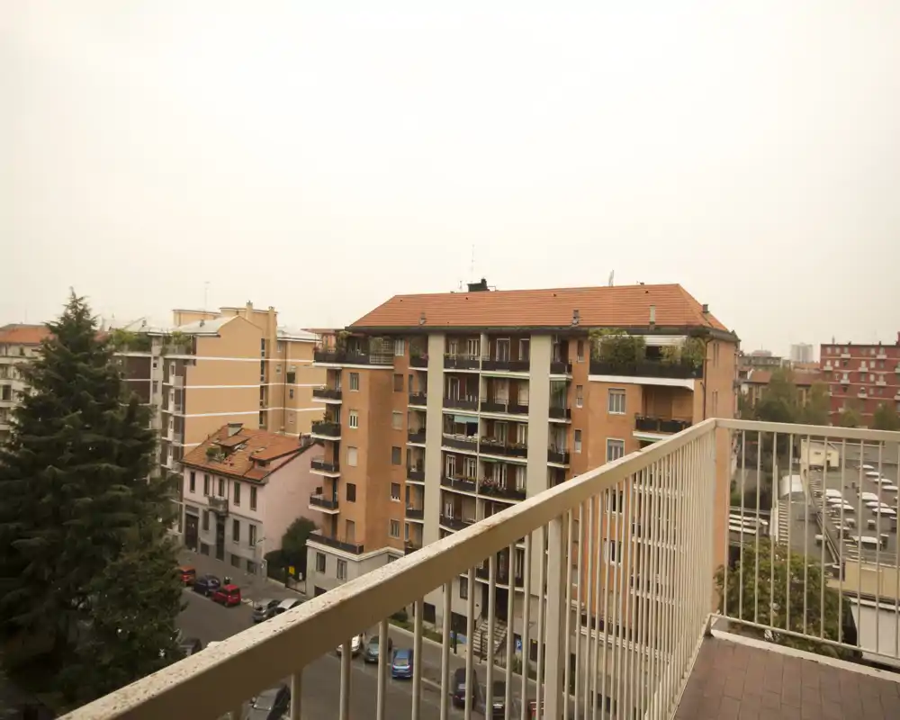Balcone con ringhiera in primo piano che si affaccia su palazzi residenziali e una via alberata; vista urbana ampia.