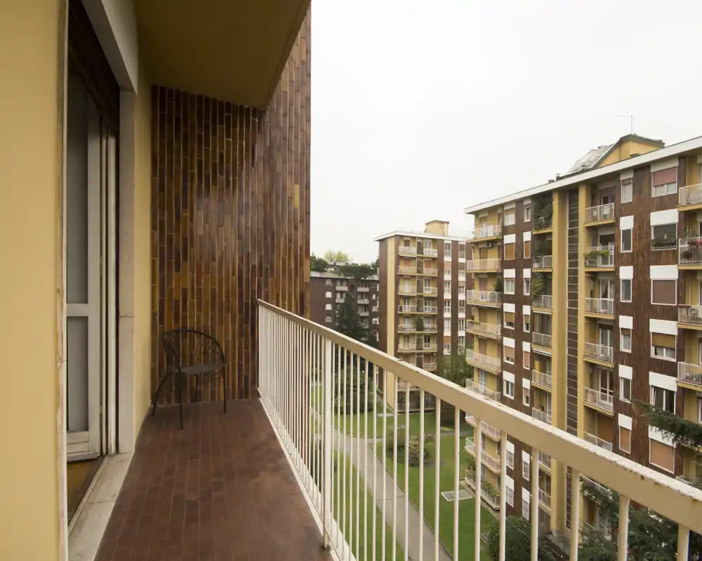 Balcone soleggiato con pavimento piastrellato e ringhiera metallica che dà su un cortile e palazzi vicini; piccola sedia e accesso diretto dall'appartamento.