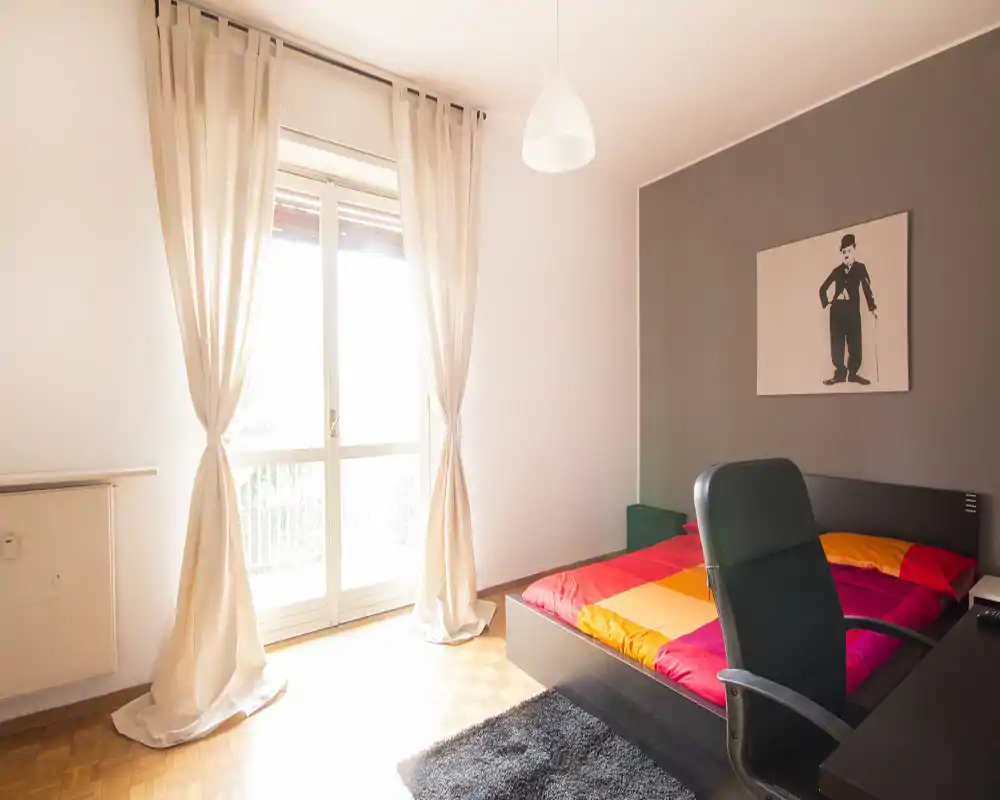 Camera da letto moderna e luminosa con ampia finestra e biancheria colorata. La stanza dispone di un letto matrimoniale, scrivania con sedia, tappeto e quadro decorativo, perfetta per un annuncio in affitto.