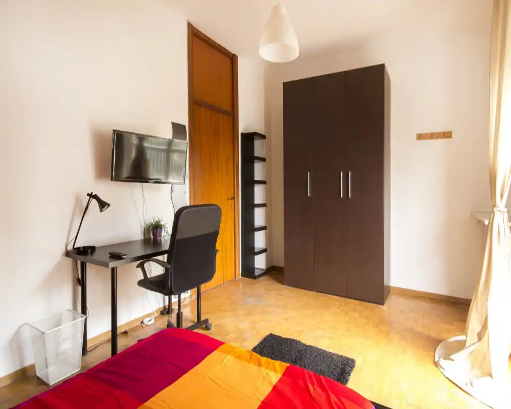Camera da letto luminosa e semplicemente arredata con copriletto colorato, scrivania e armadio. La luce naturale dalla finestra crea un'atmosfera calda e ordinata, ideale per affitti brevi.
