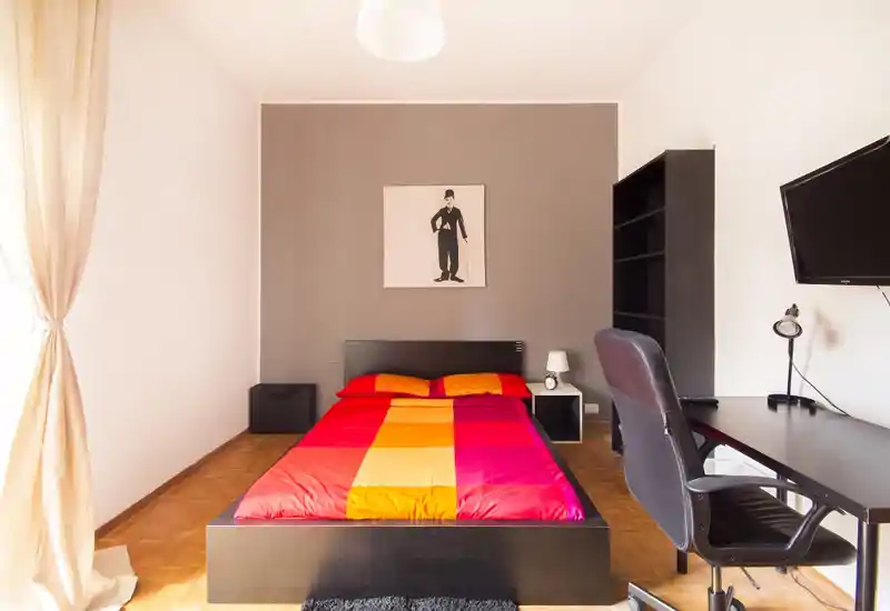 Camera da letto luminosa e moderna con letto matrimoniale al centro con biancheria rossa e arancione, scrivania e sedia a destra, libreria e comodino con lampada. Ampia finestra con tende chiare che illumina naturalmente la stanza, con un quadro minimal sulla parete d'accento.