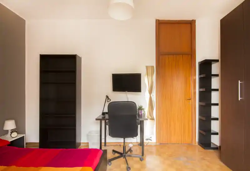 Camera da letto luminosa e ben organizzata con letto colorato, scrivania e scaffali. L'ambiente dispone di un'area lavoro con TV a parete e pavimento in legno, ideale per riposare o lavorare da remoto.