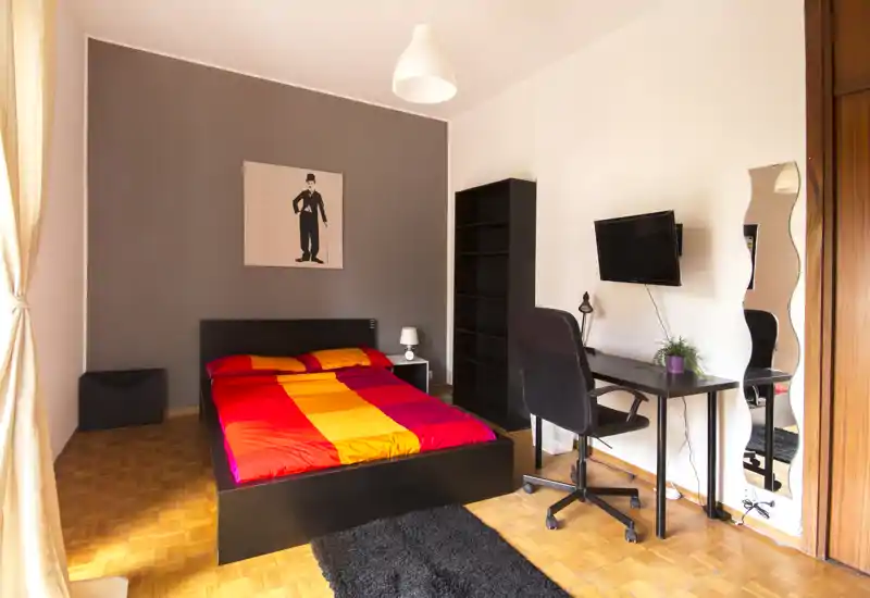 Camera da letto moderna e luminosa con letto matrimoniale con copriletto rosso/arancione, scrivania con sedia e TV a parete, libreria alta e specchio a figura intera — perfetta per una persona o coppia.