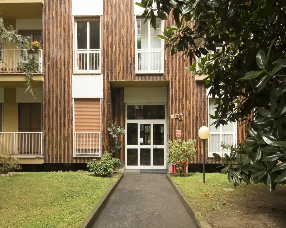 Ingresso principale di un condominio con vialetto asfaltato, porta d'ingresso in vetro a doppia anta e area verde con prato e cespugli, incorniciato da alberi.