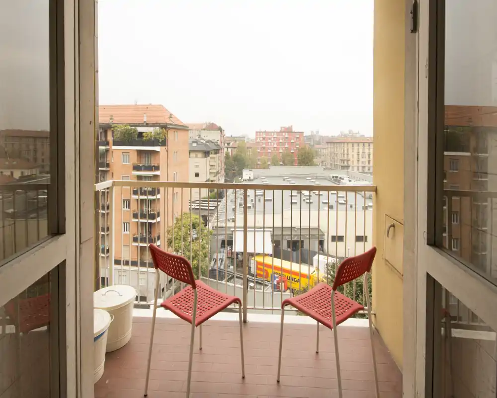 Piccolo balcone urbano con due sedie rosse che si affacciano sui palazzi vicini — ideale per un caffè mattutino con vista sulla città.