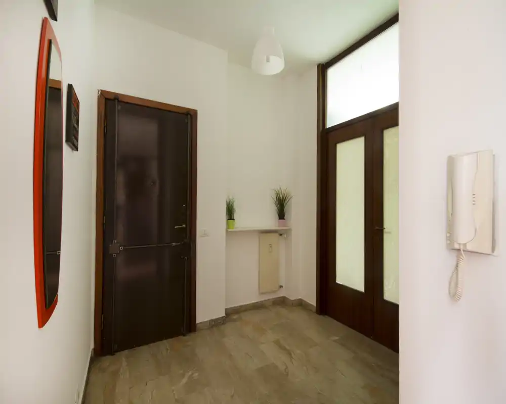 Ingresso luminoso e pulito con porta principale robusta, porta secondaria vetrata, citofono a parete e decorazioni semplici — ideale come spazio d'ingresso accogliente.