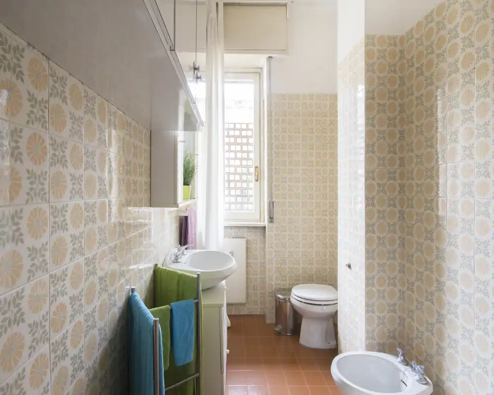 Bagno luminoso in stile vintage con piastrelle decorative, lavabo, WC e bidet. La luce naturale dalla finestra illumina le piastrelle del pavimento color terracotta.