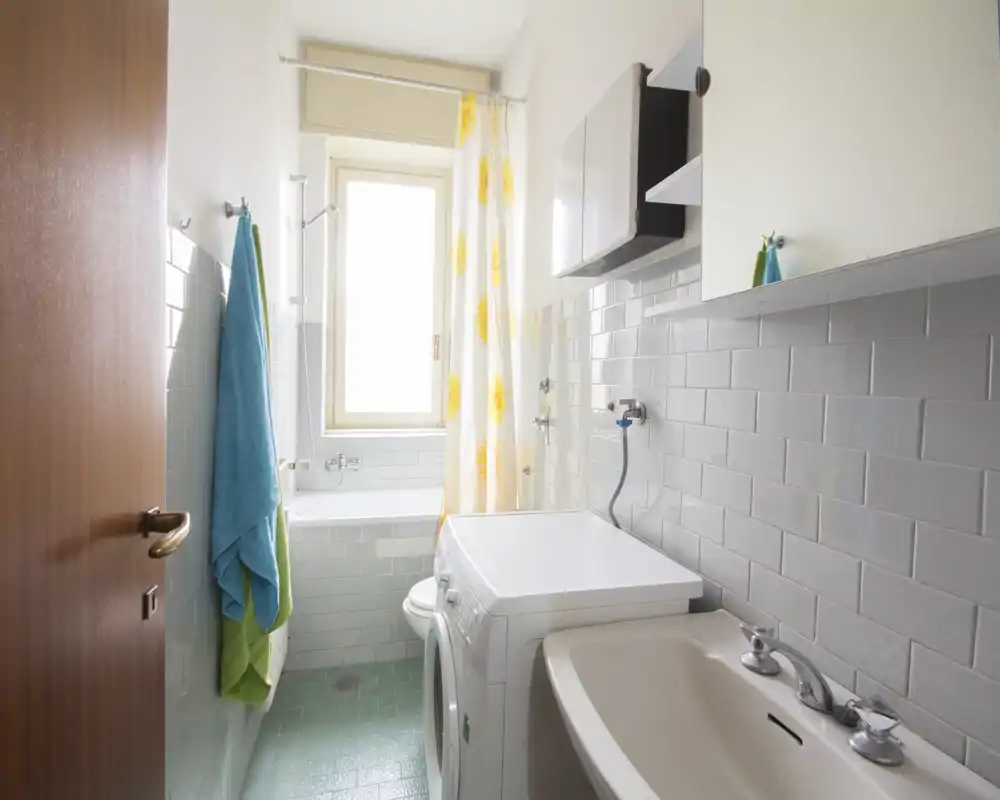 Bagno pulito e luminoso con vasca, lavabo e lavatrice; pareti piastrellate bianche e una finestra soleggiata creano un’atmosfera ariosa.