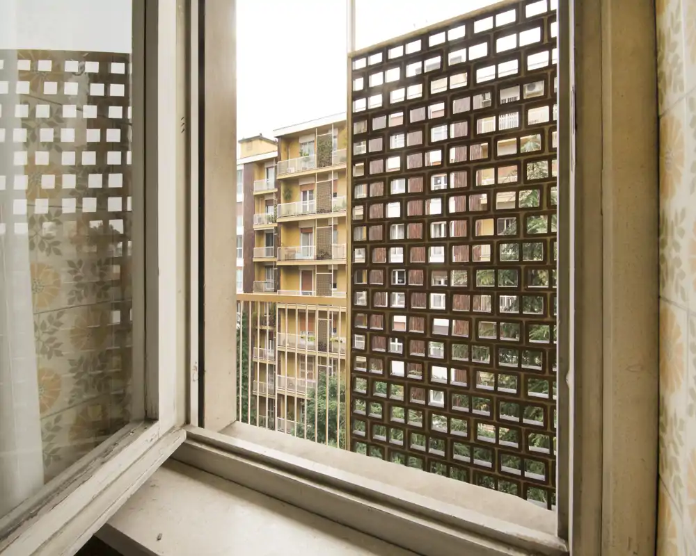 Finestra aperta con affaccio su un piccolo balcone/parete metallica schermante e palazzi vicini; vista urbana dai toni caldi.