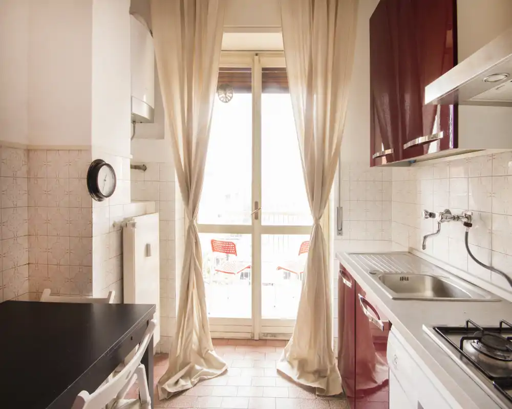 Cucina compatta e luminosa con mobili rosso bordeaux, lavello in acciaio e un piccolo tavolo da pranzo accanto alla porta sul balcone con tende leggere — perfetta per un appartamento in città.