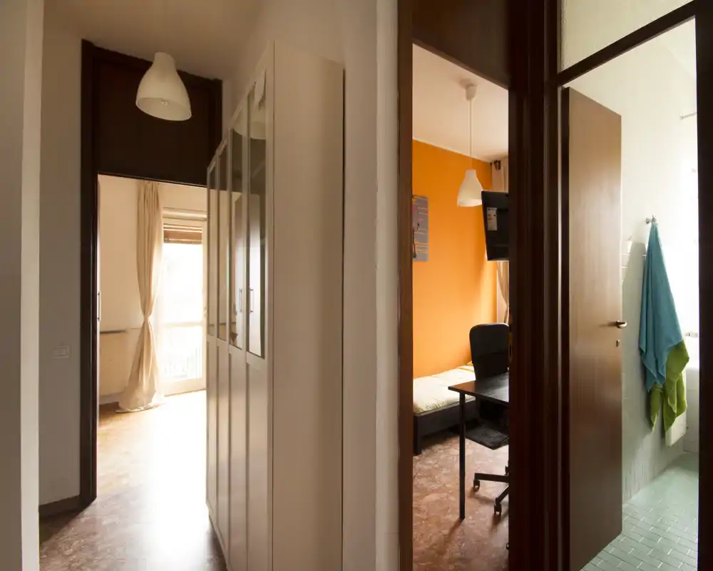 Camera da letto compatta visibile attraverso l’anta di una porta con parete arancione, singolo letto e una scrivania con sedia — luce naturale proveniente da una finestra vicina.