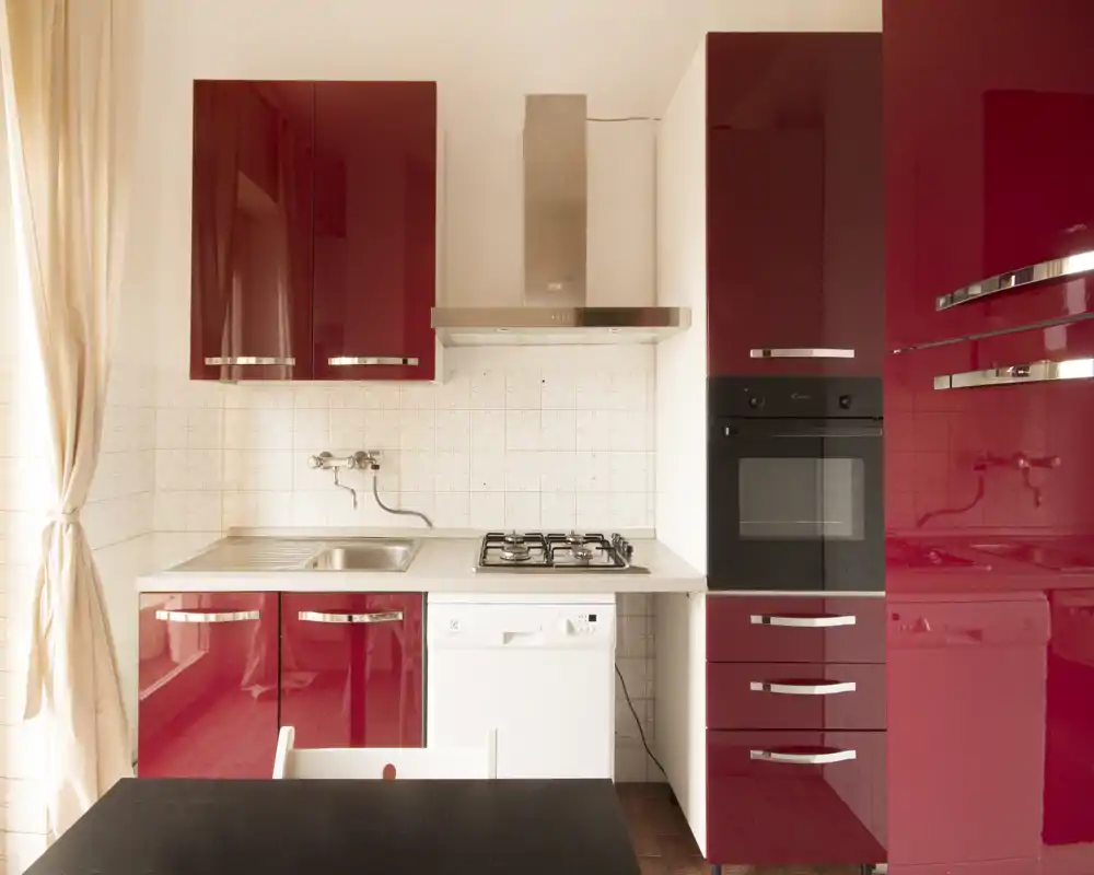 Cucina compatta moderna con mobili rossi lucidi, lavello in acciaio e piano cottura a gas; luce naturale intensa e piccolo piano pranzo, pratica per l'uso quotidiano.